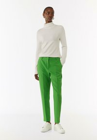 Weißer Rollkragenpullover, figurbetont mit langen Ärmeln; leuchtend grüne maßgeschneiderte Hose; weiße Sneakers; glatte Textur und moderner Schnitt.