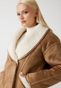 Giacca in suede marrone con colletto in pelliccia sintetica crema, grandi tasche e vestibilità morbida. Gli accenti includono chiusure con bottoni e dettagli di cucitura.
