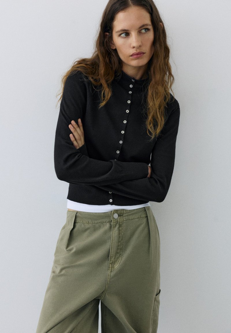 Femme aux longs cheveux ondulés porte un cardigan noir boutonné et un pantalon vert olive, debout les bras croisés devant un fond clair uni.
