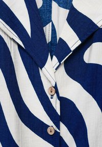 Button-up shirt featuring a large navy and white wavy pattern, with a fold-down collar and a wooden button detail. Textured fabric.