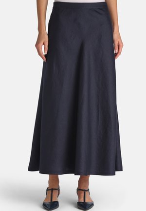 Frau trägt einen marineblauen, hoch taillierten Midi-Rock in A-Linien-Form, kombiniert mit schwarzen T-Riemchen-Pumps mit spitzen Zehen vor neutralem Hintergrund.