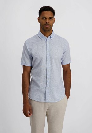 Man draagt een lichtblauw overhemd met korte mouwen, met een subtiel bladpatroon en beige broek, staand tegen een effen achtergrond.