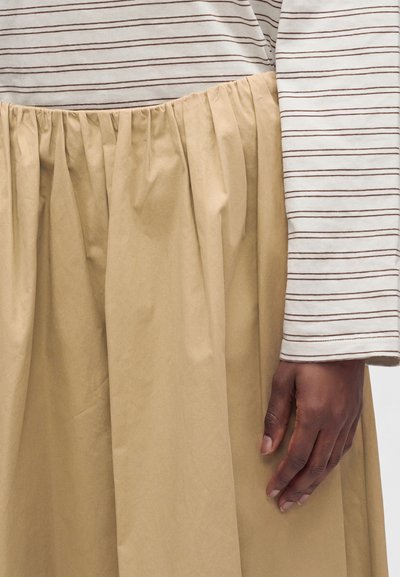 Main droite d'une personne portant une chemise blanche à manches longues avec de fines rayures marron et une jupe ou un pantalon plissé beige.