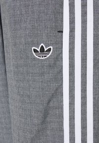 Pantalones deportivos grises con un patrón tejido texturizado, rayas laterales blancas y un logotipo bordado en blanco y negro.