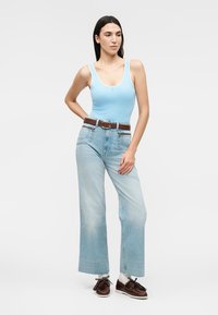Femme debout portant un débardeur bleu clair, un jean taille haute à jambes larges, une ceinture marron, des chaussettes blanches et des chaussures en cuir marron.
