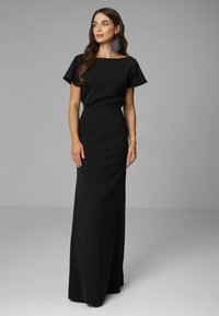 Czarna sukienka do ziemi ma krótkie, falujące rękawy, dekolt łódkowy oraz dopasowaną sylwetkę wykonaną z gładkiego materiału.