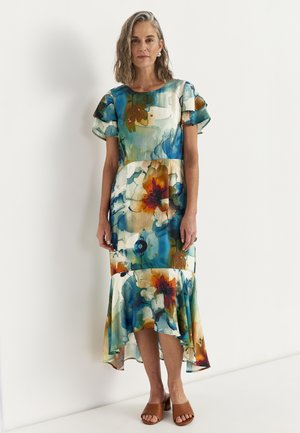 Kvinde stående iført en kortærmet, asymmetrisk blomstret kjole med mønstre i blå, orange og grønne akvarelfarver, kombineret med brune åbne tå-sandaler.
