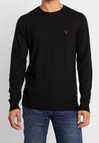 Black knit sweater with a crew neck, ribbed cuffs and hem, featuring a small embroidered logo on the chest, paired with blue jeans.