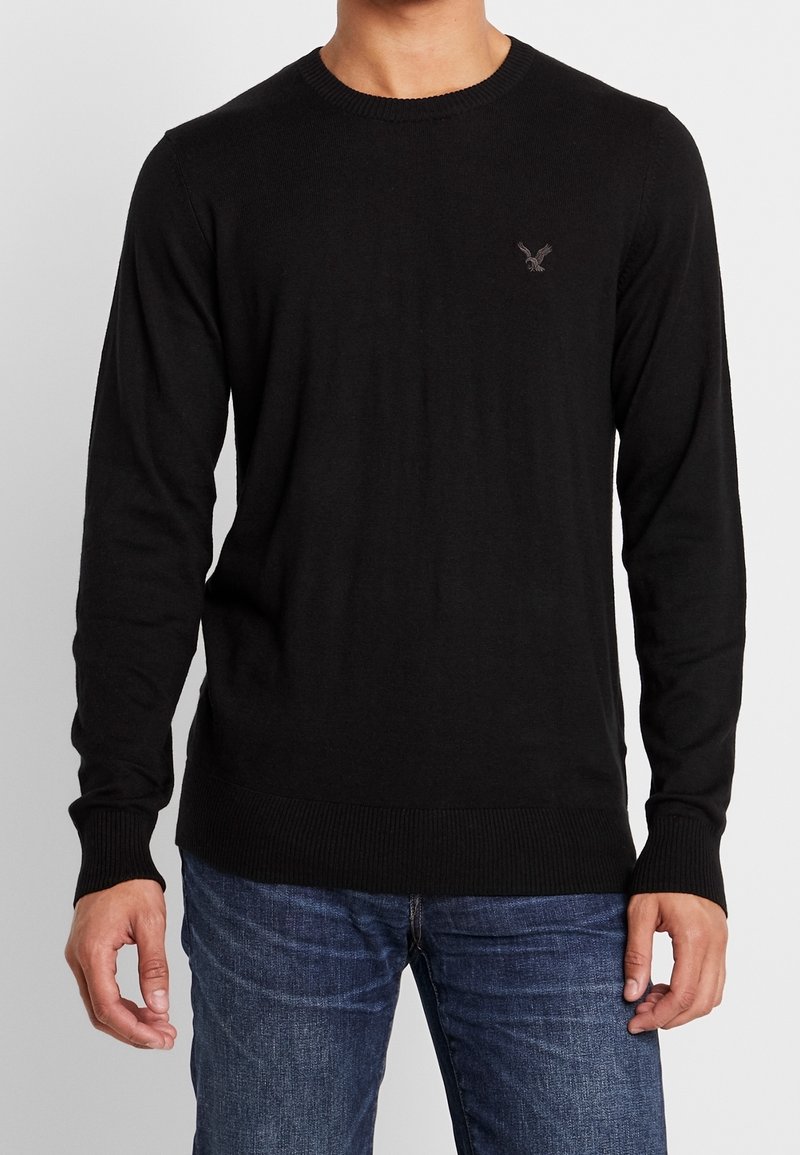 Black knit sweater with a crew neck, ribbed cuffs and hem, featuring a small embroidered logo on the chest, paired with blue jeans.