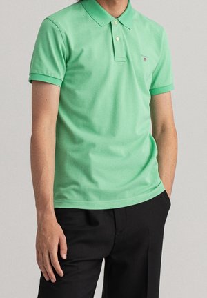 Hombre que lleva una camisa polo de manga corta de color verde claro y pantalones negros con una mano en el bolsillo sobre un fondo liso.