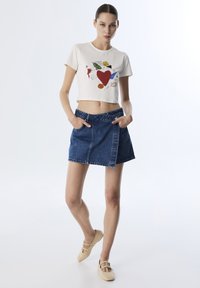 Camiseta corta color crema con un gráfico colorido, combinada con una mini falda de mezclilla azul que cuenta con un bolsillo delantero y trabillas para cinturón. Calzado ligero.