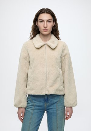Ung person med langt hår iført en beige, blød lynlåstrøje og blå jeans, står foran en ensfarvet hvid baggrund.