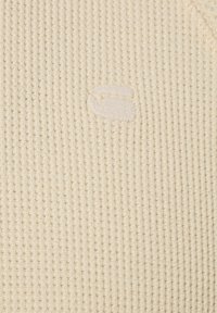 Beiger, gerippter Stoff mit einem dezenten, gestickten Logo. Die Textur ist weich und besitzt ein atmungsaktives Muster. Keine auffälligen Hardware-Elemente oder Akzente sichtbar.