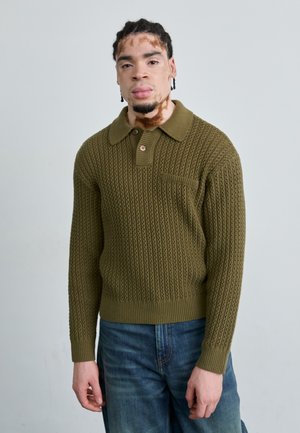 Knit pullover in olive green with a textured pattern, collar, and button detail. Fitted silhouette with ribbed cuffs and hem. Jeans visible.