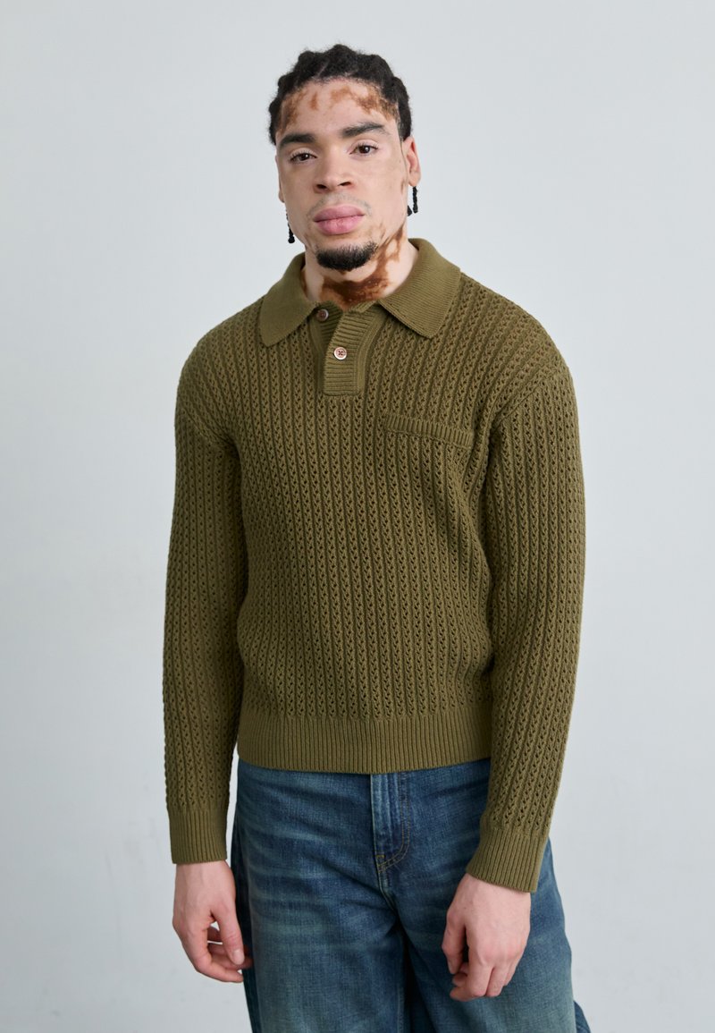 Knit pullover in olive green with a textured pattern, collar, and button detail. Fitted silhouette with ribbed cuffs and hem. Jeans visible.