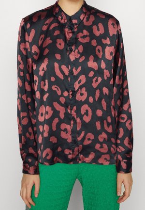 Chemisier en satin noir avec un imprimé léopard rouge, présentant un col, un devant à boutons et des manches longues. Porté avec un pantalon vert texturé.