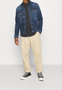 Hombre que lleva una chaqueta de denim azul oscuro, una camisa negra de botones, pantalones beige arremangados y zapatillas altas negras, de pie contra un fondo liso.