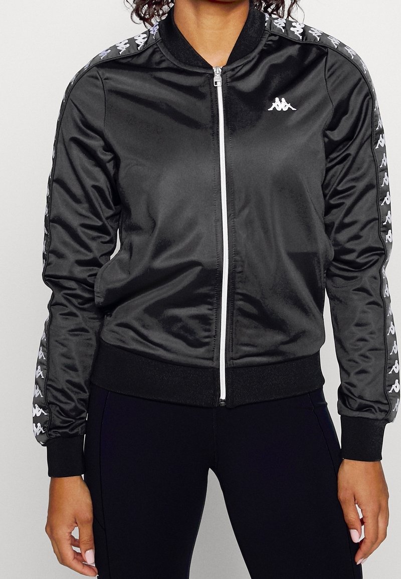 Femme portant une veste zippée en satin noir avec des logos blancs sur les épaules et les manches, assortie à un pantalon noir ajusté.