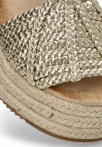 Correa en tono dorado tejido con textura trenzada, montada sobre una suela de plataforma de cuerda natural; detalle de plantilla con textura de madera circular.