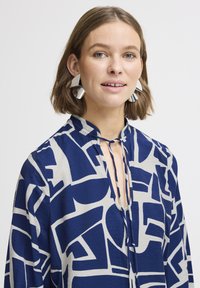 Blusa blu navy con motivi bianchi astratti, maniche lunghe e dettaglio del colletto con fiocco. Il modello indossa grandi orecchini geometrici in argento.