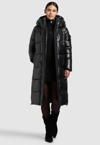 Crni dugački kaput s podstavom i kapuljačom, sjajnog završnog sloja, s patentnim zatvaračem sprijeda i velikim prošivenim dijelovima. Nošen preko crne pletene haljine i crne obuće.