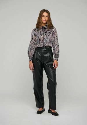 Γυναίκα φορώντας μια μπλούζα με πατέντα paisley μακρυμάνικη, μαύρο δερμάτινο παντελόνι με ψηλή μέση και μαύρα φλατ παπούτσια, στέκεται μπροστά σε μια μονόχρωμη φόντο.