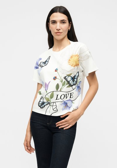 Γυναίκα που φοράει λευκό κοντομάνικο πουκάμισο με κέντημα λουλουδιών και πεταλούδας και την λέξη «LOVE», συνδυασμένο με σκούρο παντελόνι, στέκεται μπροστά σε λευκό φόντο.