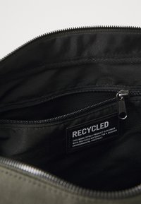 Melnas somas iekšpuses, kas aprīkota ar rāvējslēdzēju, ar etiķeti, uz kuras rakstīts "RECYCLED" uz auduma uzlīmes. Redzama faktūra un metāla piederumi.