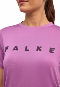 Jasnofioletowa sportowa koszulka z teksturowanej tkaniny, z wyraźnymi czarnymi literami piszącymi "FALKE" z przodu. Krótkie rękawy i okrągły dekolt.