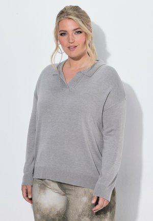 Blonde vrouw met vastgebonden haar draagt een lichtgrijze trui met lange mouwen en een broek met patroon, staand voor een effen witte achtergrond.