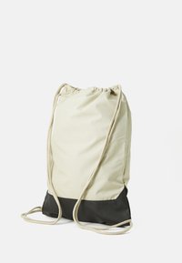 Sac à dos à cordon de couleur beige clair avec un fond noir uni. Fabriqué en tissu durable et équipé de cordons de contraste pour un transport facile.