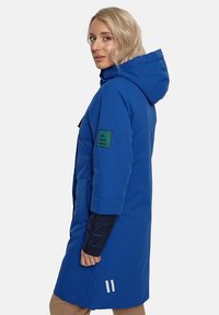 Zilgs parka ar kapuci, garš dizains, sānu kabatas, zaļa zīmola uzlīme uz piedurknes un balti akcenti tuvu apakšmalai. Gluda tekstūra.