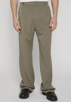 Muž nosiaci voľné khaki nohavice a čierne topánky, stojaci pred jednoduchým svetlým pozadím.