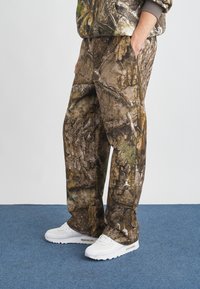 Παντελόνι καμουφλάζ με υφή σε καφέ και πράσινο χρώμα που θυμίζει φλοιό και φύλλα, με πλαϊνές τσέπες και λευκή λεπτομέρεια λογότυπου.