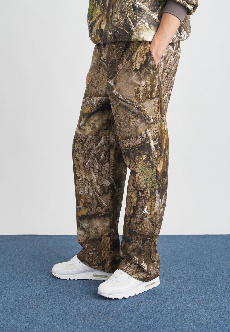 Παντελόνι καμουφλάζ με υφή σε καφέ και πράσινο χρώμα που θυμίζει φλοιό και φύλλα, με πλαϊνές τσέπες και λευκή λεπτομέρεια λογότυπου.