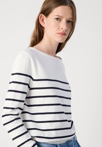 Witte lange mouwen top met marineblauwe horizontale strepen, ronde halslijn, zachte stof en klein rood logo op de borst. Combinatie met blauwe jeans.