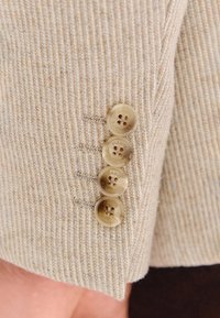 Cztery beżowe guziki przyszyte do prążkowanego beżowego rękawa, ułożone pionowo z widocznymi dziurkami na guziki i detalami szycia.