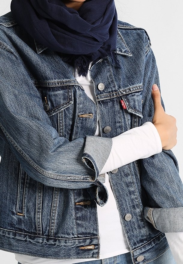Personne portant une veste en jean bleue par-dessus une chemise blanche à manches longues et une écharpe bleu foncé, les bras croisés à la taille.