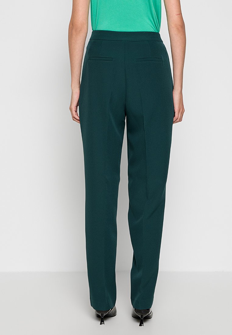 Donkergroene broeken met een recht model, een gladde textuur en twee achterzakken. Draagt een groen T-shirt met korte mouwen en zwarte schoenen.