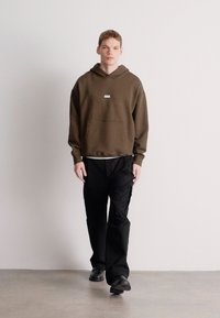 Ruda gobtuvuota sportinė palaidinė su priekyje esančia kengūros kišene ir mažu logotipu; derinama su juodomis, laisvai krentančiomis kelnėmis ir juodais batais.