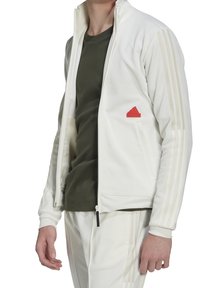 Vitt sportjacka med ljusbeiga ränder, dragkedjestängning och röd triangel-logotyp på bröstet. Matchas med en mörkgrön t-shirt.