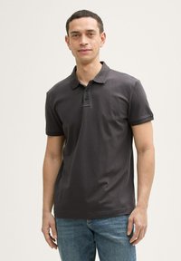 Polo shirt gris foncé à manches courtes, avec patte de boutonnage à trois boutons et texture subtile. Porté avec un jean en denim bleu. Design simple et classique.