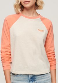 Lange mouwen baseballstijl top in crème met zalmokleurige mouwen. Bevat het "SuperDry" logo in oranje op de borst. Zachte textuur.