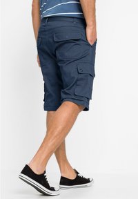 Pantaloni cargo blu navy con molte tasche, realizzati in tessuto resistente. Indossati con sneakers nere con lacci bianchi e profilo basso.