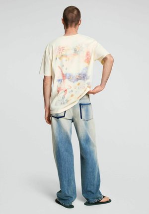 Person trägt ausgewaschene blaue Jeans mit dunkelblauen Taschenrändern und ein cremefarbenes T-Shirt mit bunten abstrakten Drucken, steht mit der Hand in der Hüfte.