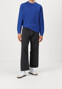 Hellblauer Pullover mit Zopfmuster und Rundhalsausschnitt, kombiniert mit locker sitzenden schwarzen Jeans und weißen Turnschuhen mit braunen Akzenten.