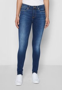 Jeans ajustados en denim azul oscuro, con cinco bolsillos, costuras en contraste y cierre de cremallera con botón. Combinados con zapatillas blancas.