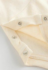 Valkoinen puuvillabodysuit, jossa on ribbireunus kauluksessa ja napit alapuolella. Pehmeä rakenne ja hienovaraiset ompeluyksityiskohdat.