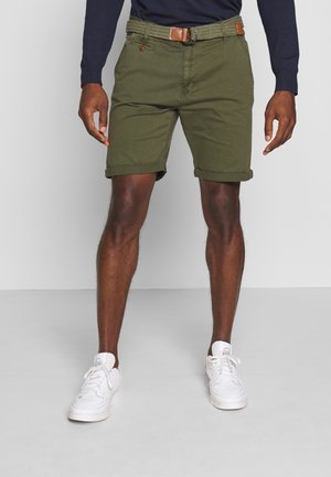 Homme portant un short olive retroussé avec une ceinture tissée, une chemise à manches longues bleu marine et des baskets blanches, debout devant un fond gris.