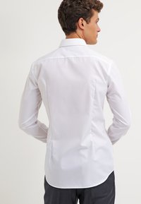 White, long-sleeve button-up shirt with a standard collar and back darts. Smooth texture; fitted design; curved hem at the back.
